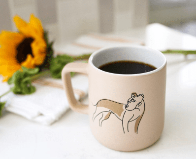 close up of a cup of coffee with an illustration of a pitup in a line art style sitting on a desk with a sunflower in the background and blurry